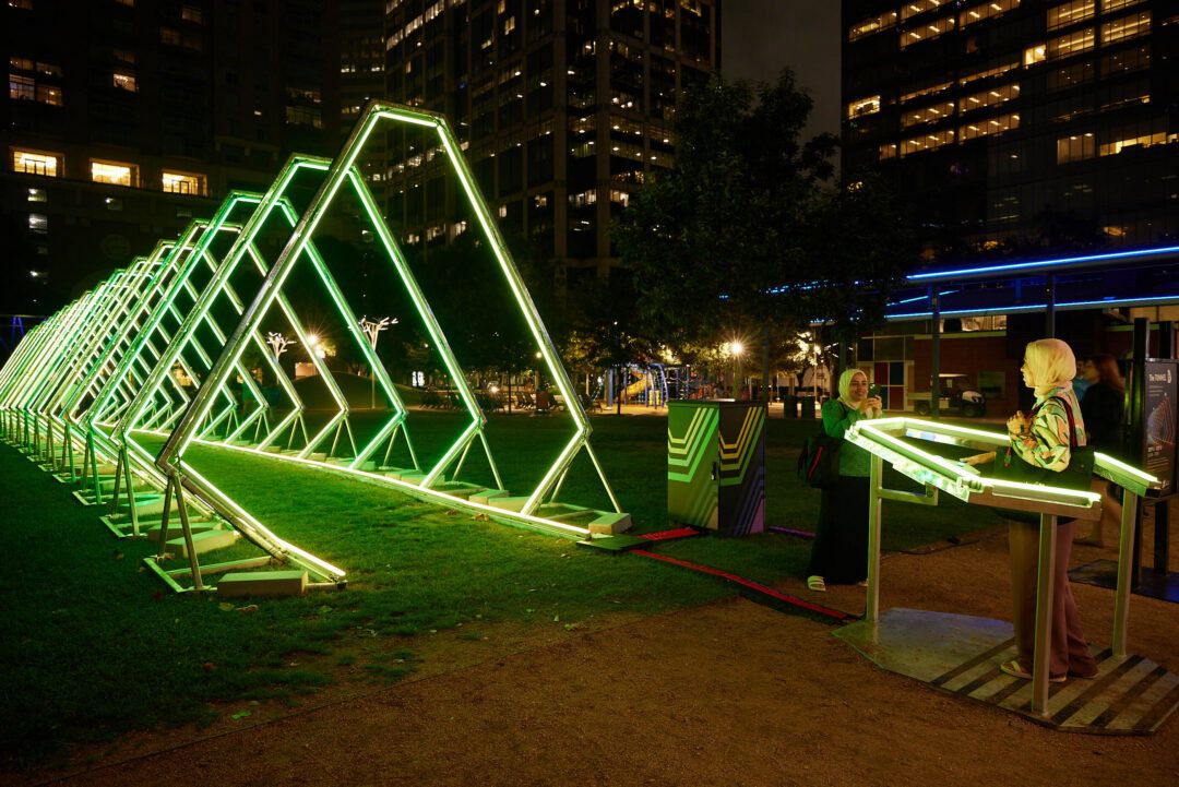 A pair of people enjoy an installation piece that's shaped like a tunnel and lit up green. One person is photographing the other standing inside a console and pushing buttons.