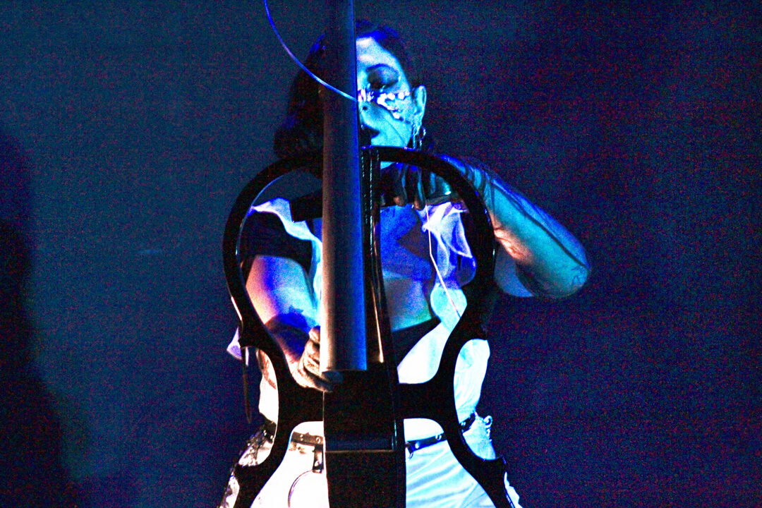 A musician in blue light playing cello and looking directly at the viewer.