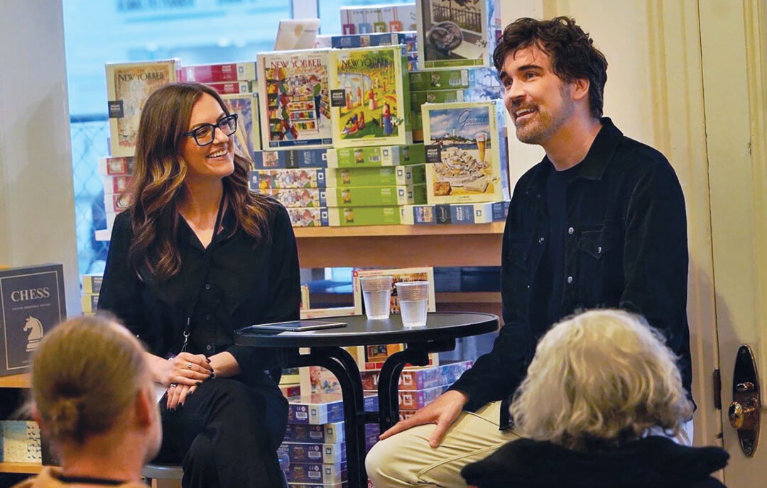A femme-presenting person and a masculine-presenting person talk in front of a window full of puzzles to an audience.