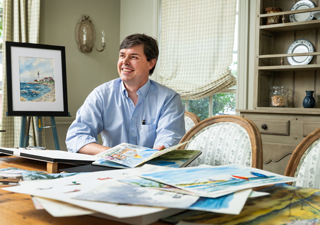 A masculine-presenting person smiling at a table full of watercolor paintings.
