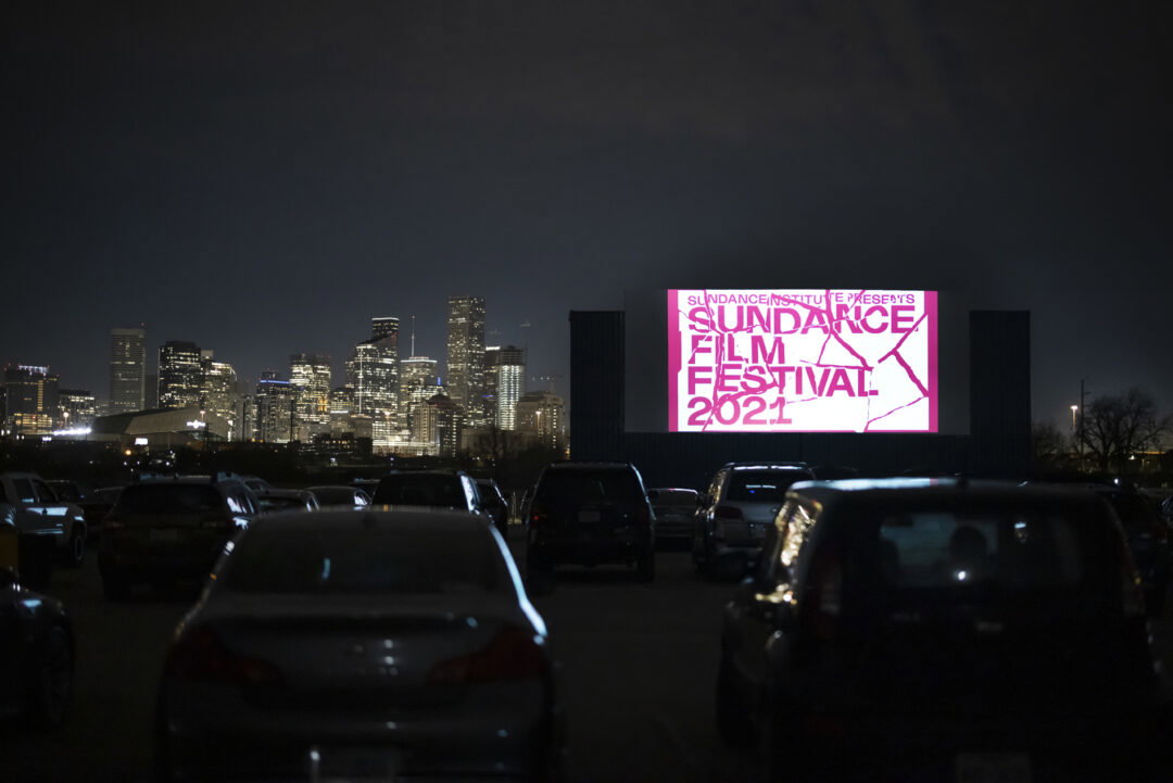 A drive-in movie theater screen is playing Sundance Festival favorites from 2021 in front of lines of cars. The Houston skyline is lit up in the background.