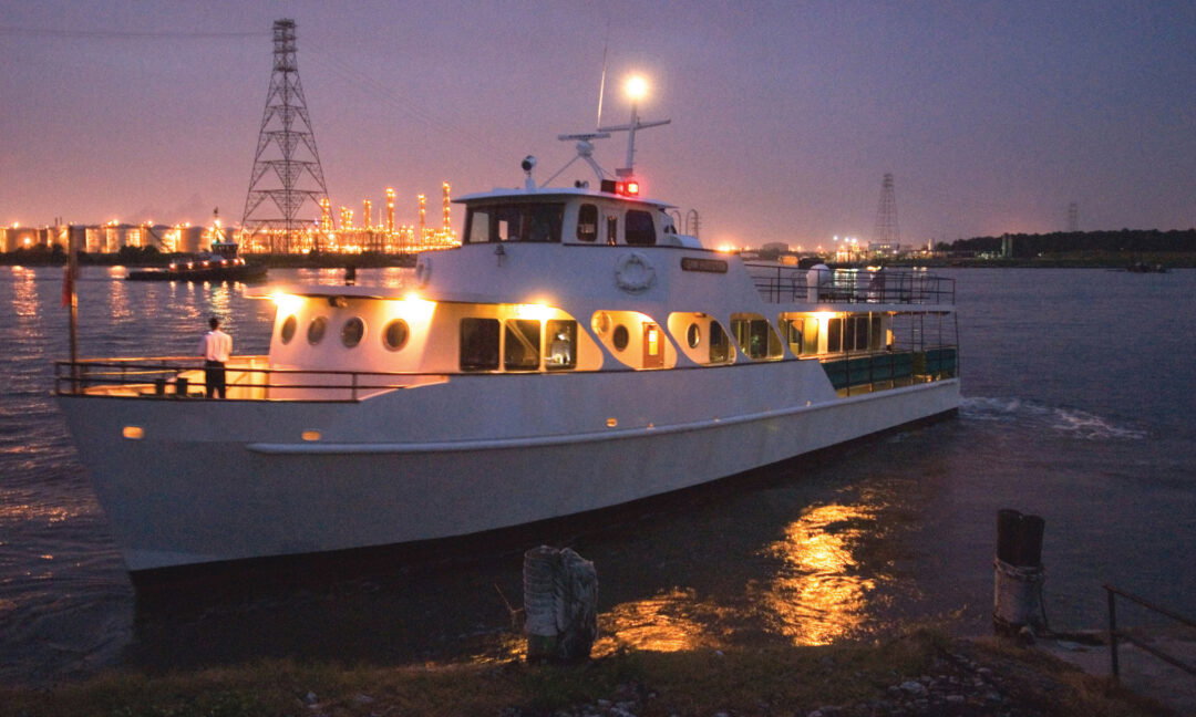 A midcentury modern passenger boat lit up at night. A person stands on the deck.