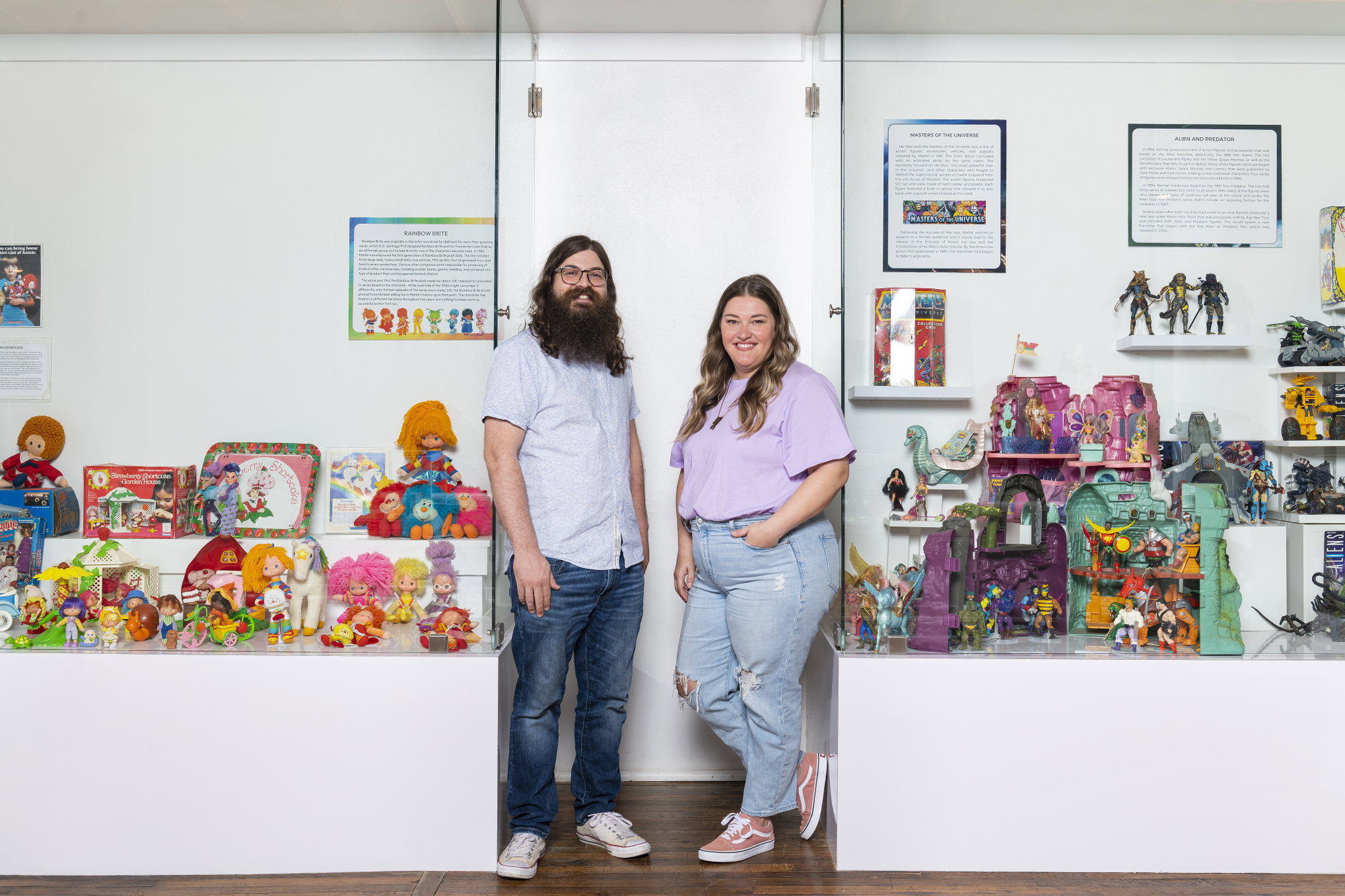 Two smiling people pose in front of a museum display full of toys.